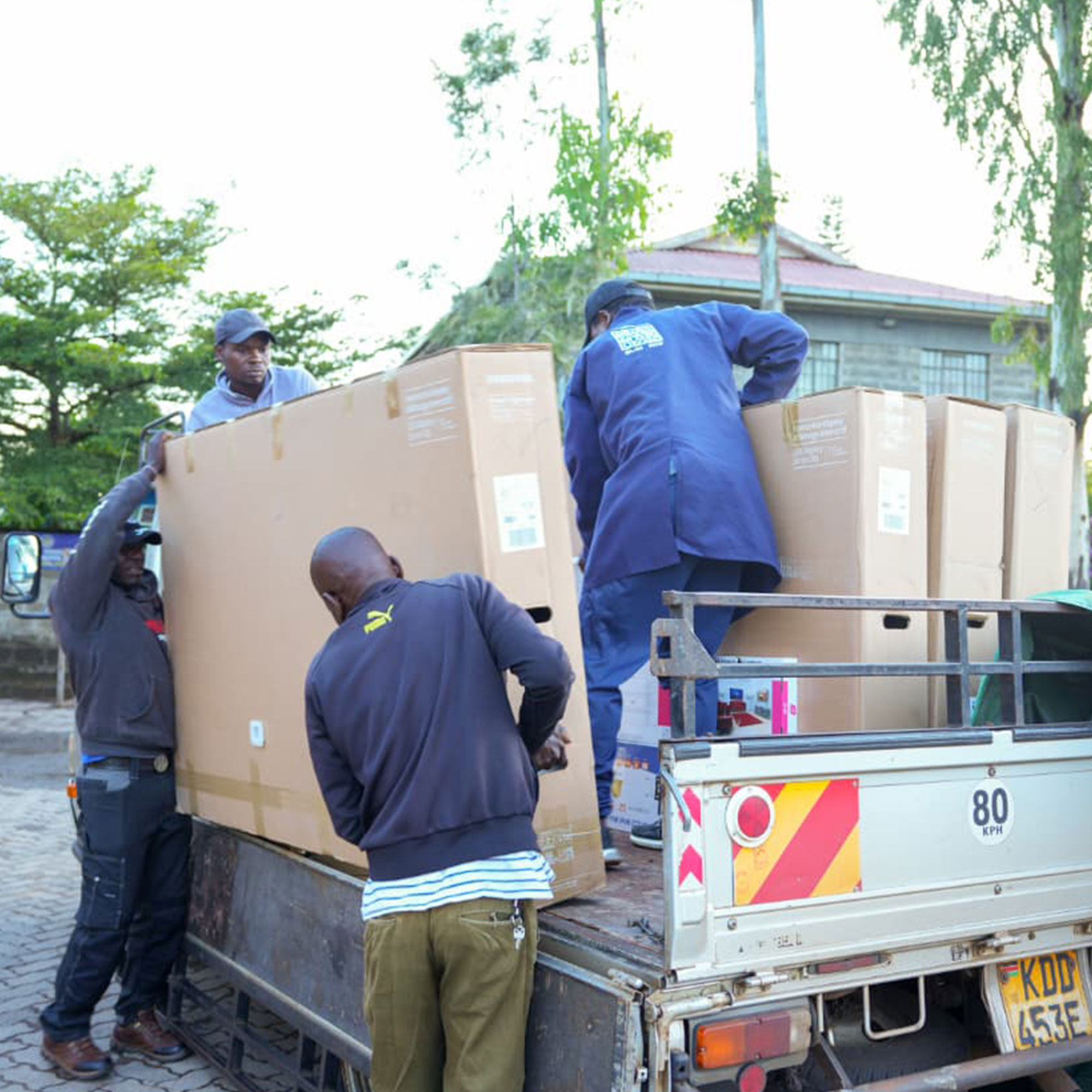 Workers in blue uniforms load large cardboard boxes onto a flatbed truck outdoors.