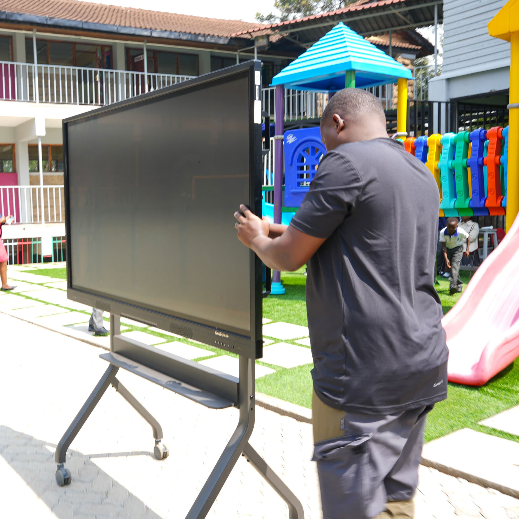 Man in a dark shirt carries a large flat-screen TV on a wheeled stand at an outdoor playground.