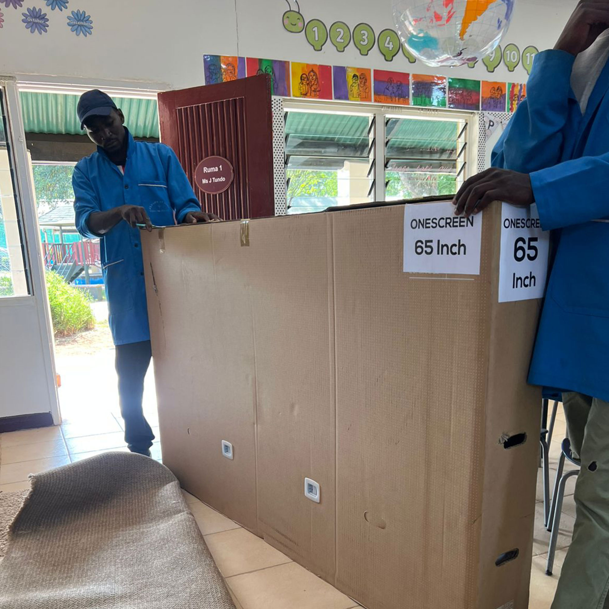 Two workers in blue uniforms seal a large cardboard box labeled '65 Inch' inside a room, preparing a TV packaging shipment.