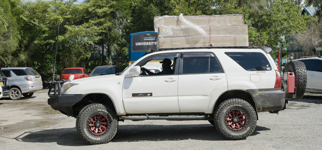 White SUV with a large pallet load strapped to the roof, parked in a lot, off-road tires with red rims visible on the side.