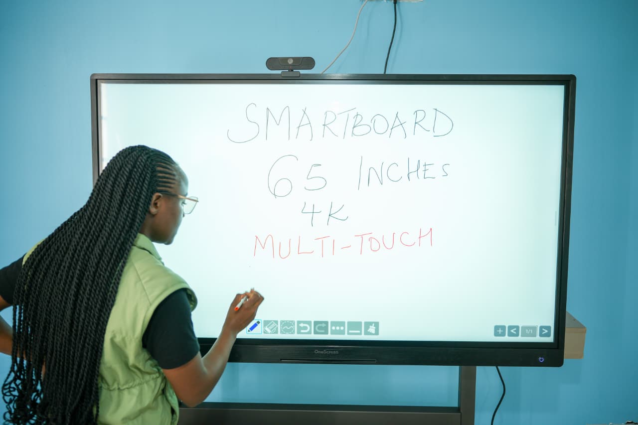 Person standing at a 65-inch, 4K multi-touch Smartboard on a blue wall, writing text that reads 'SMARTBOARD 65 INCHES 4K MULTI-TOUCH' at the board's surface.